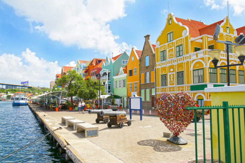 Curaçao Bunte Häuserfassaden im niederländischen Kolonialstil an der Uferpromenade von Willemstad, Curaçao. Im Vordergrund ein Herz aus Liebesschlössern, alte Kanonen und Sitzbänke am Wasser. Menschen sitzen in Cafés unter schattigen Bäumen, ein Ausflugsboot lieg