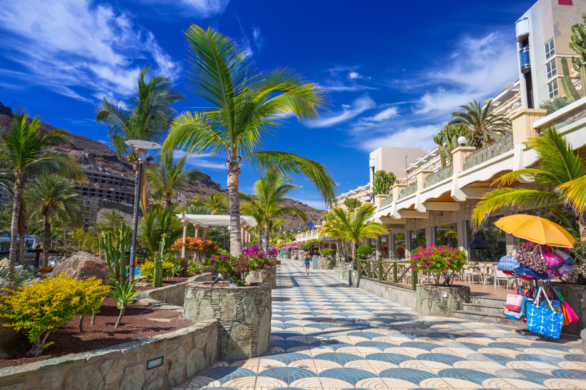 Promenade in Taurito auf Gran Canaria mit Palmen, Blumenbeeten und Gebäuden unter blauem Himmel