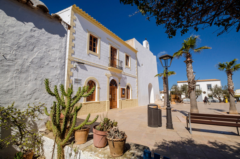Sonnige Plaza mit weißen Gebäuden im mediterranen Stil in Sant Francesc Xavier, Formentera. Vor dem Hauptgebäude stehen Palmen, ein Kaktus in einem Blumentopf und eine Bank, während eine Person auf einem Fahrrad vorbeifährt.