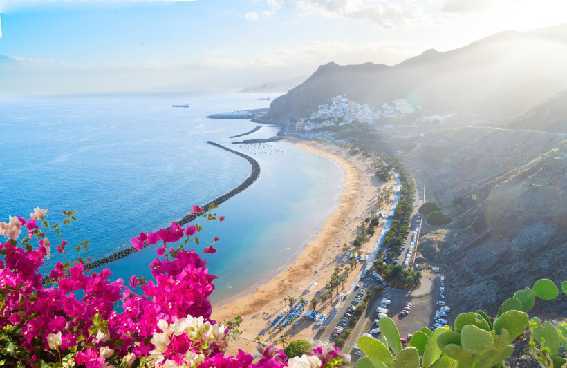 Blick auf Playa de Las Teresitas auf Teneriffa mit goldenem Sand, türkisblauem Meer, Bergen und bunten Bougainvillea-Blüten im Vordergrund.