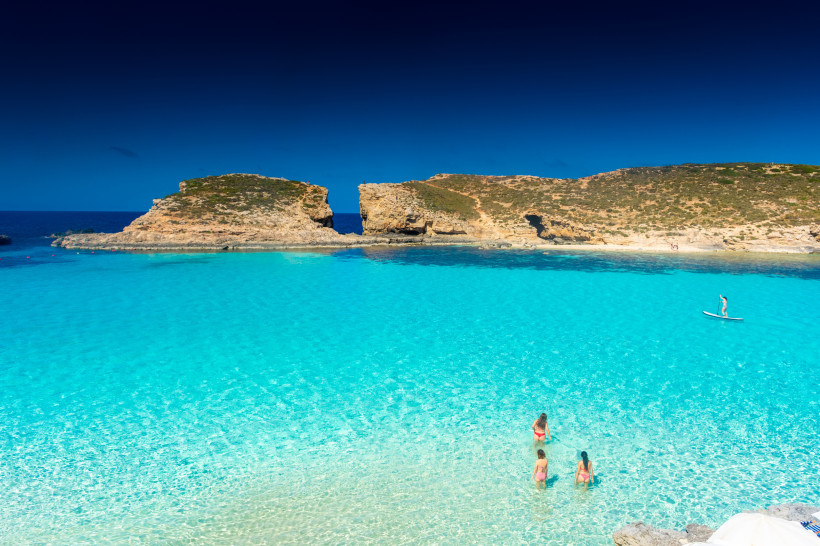 Kristallklares türkisblaues Wasser der Blue Lagoon auf Comino, Malta, mit Badegästen und Paddleboarder unter blauem Himmel