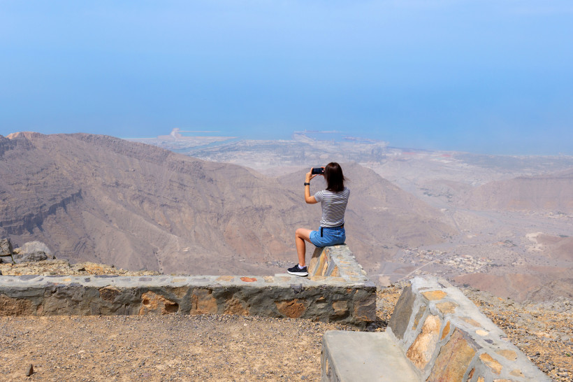Frau fotografiert die Aussicht vom Jebel Jais auf Berge, Wüste und Meer in Ras Al Khaimah – beliebter Aussichtspunkt im Hadschar-Gebirge.