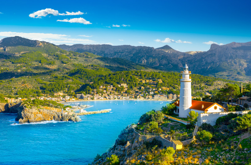 Mallorca - Port de Sóller Eine wunderschöne Hafenbucht auf Mallorca mit klarem türkisfarbenem Wasser. Segelboote und Yachten liegen im Hafenbecken, während die umliegenden Gebäude und die grüne Landschaft im Hintergrund zu sehen sind. Im Vordergrund ist ein Leuchtturm zu erkennen.