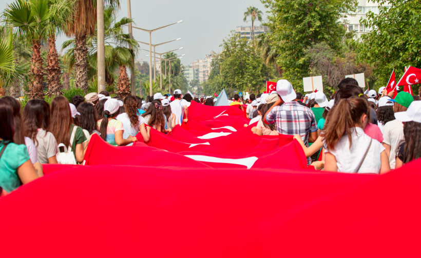 Eine große Menschenmenge, überwiegend junge Menschen, trägt eine riesige türkische Flagge bei einem Umzug durch eine palmengesäumte Straße in einer Stadt.