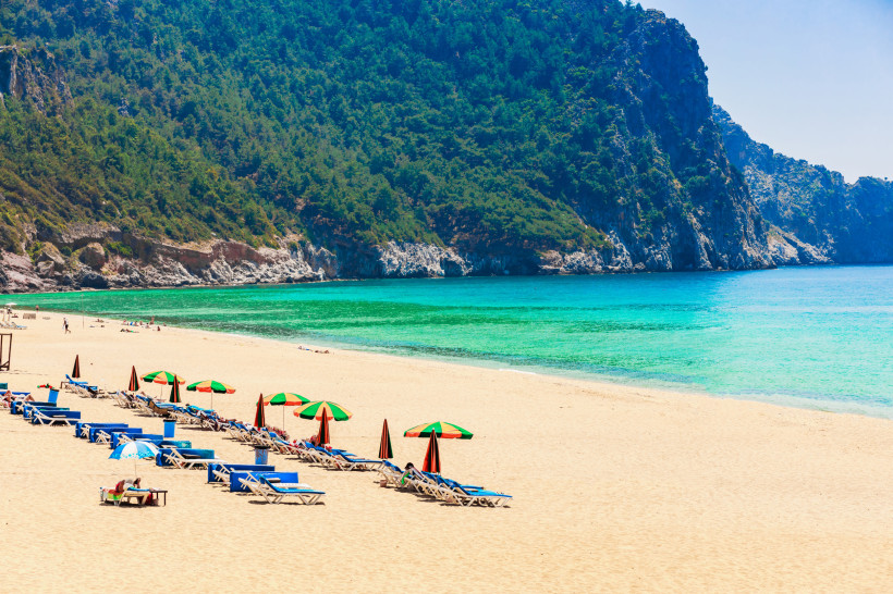 Kleopatra-Strand in Alanya mit Reihen von Sonnenliegen und bunten Sonnenschirmen vor bewaldeter Felsenküste und türkisfarbenem Meer