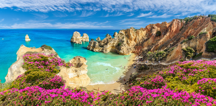 Felsige Küste der Algarve mit türkisblauem Wasser, rosa Blumen und Sandstrand zwischen Klippen
