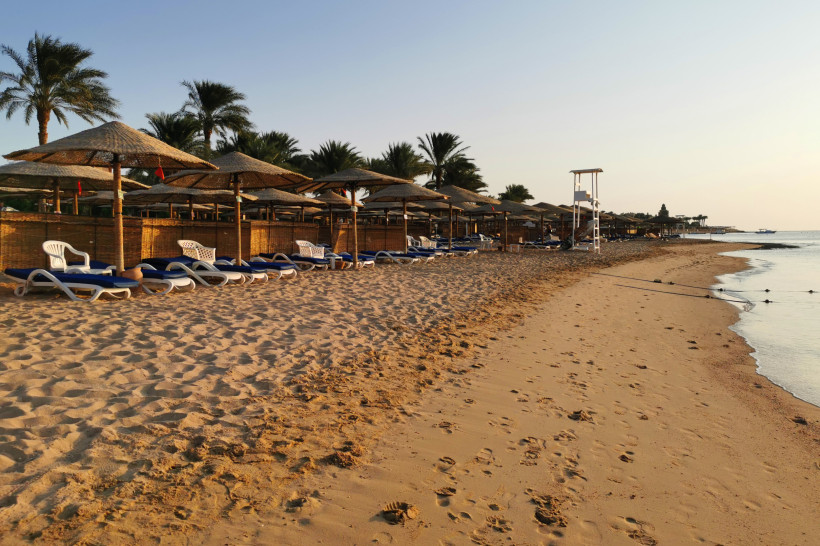 Ägypten - Makadi Bay Ruhiger Sandstrand mit Sonnenliegen unter Strohschirmen, daneben Palmen. Im Hintergrund das ruhige Meer, rechts eine weiße Rettungssitzbank. Der Himmel ist klar.