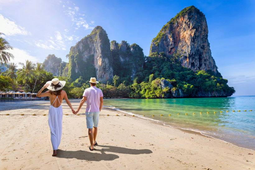Ein Paar geht Hand in Hand am tropischen Strand entlang. Im Hintergrund ragen grüne Kalksteinfelsen in den Himmel, das Meer schimmert türkis, Palmen säumen den Strand. Die Szene wirkt friedlich und romantisch. ( Railay Beach in Krabi )