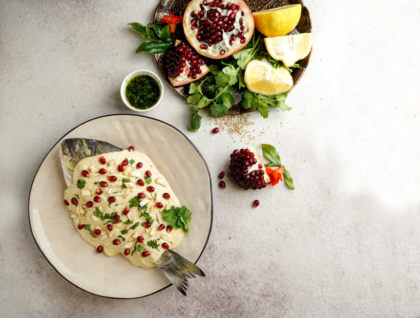 Ägypten - Makadi Bay Ganzer Fisch auf einem Teller, überzogen mit heller Sauce, bestreut mit Granatapfelkernen, Mandeln und Koriander. Daneben liegen Granatapfelhälften, Zitronenstücke und frische Kräuter.