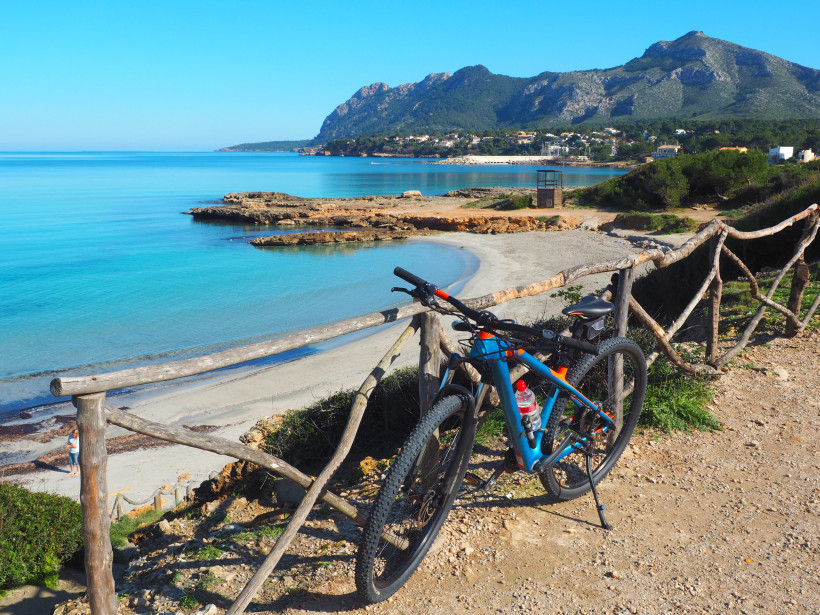 Mountainbike an einem Küstenweg oberhalb der Bucht von Alcudia mit Blick auf türkisfarbenes Meer und flache Sandbucht – ideale Bedingungen für aktiven Erholungsurlaub.