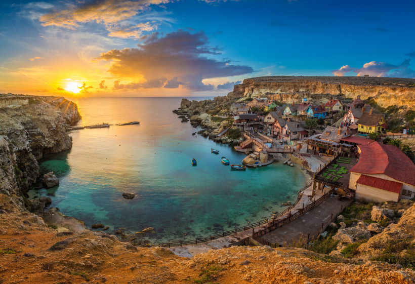 Popeye Village Malta – Filmkulisse in der Anchor Bay bei Sonnenuntergang Popeye Village in der Anchor Bay auf Malta bei goldenem Sonnenuntergang, umgeben von türkisblauem Meer und Felsenküste