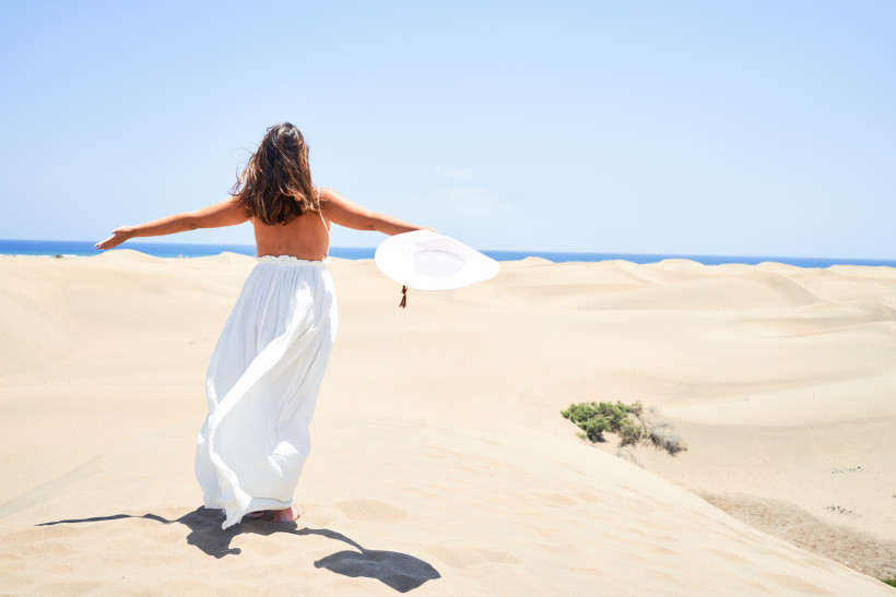 Person in weißem Kleid mit ausgebreiteten Armen auf den Dunas de Maspalomas nahe Meloneras, im Hintergrund das Meer