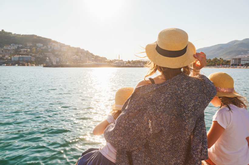 Mutter und zwei Kinder mit Strohhüten sitzen am Meer und blicken auf eine Küstenstadt im warmen Abendlicht