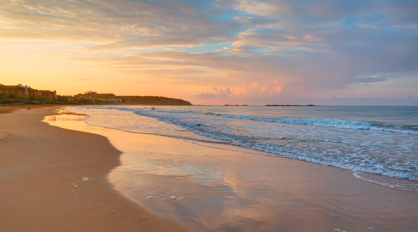 Auf dem Bild ist ein leerer, breiter Sandstrand bei Sonnenuntergang zu sehen. Das warme Licht spiegelt sich im nassen Sand wider. Die Wellen rollen ruhig ans Ufer. Links erkennt man einige Hotelgebäude in der Ferne. Der Himmel ist von zarten Wolken durchz