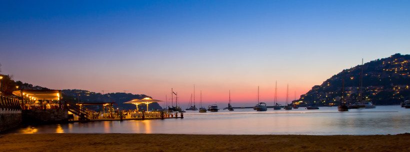 Abendlicher Blick auf den Hafen von Port d’Andratx mit vor Anker liegenden Segelbooten und beleuchtetem Steg