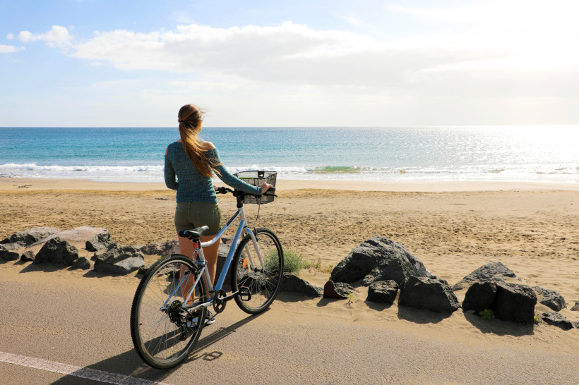 Playa Honda Lanzarote – Radfahren entlang der Strandpromenade mit Meerblick Frau mit Fahrrad an der Strandpromenade von Playa Honda auf Lanzarote mit Blick auf den goldenen Sandstrand und das türkisfarbene Meer.