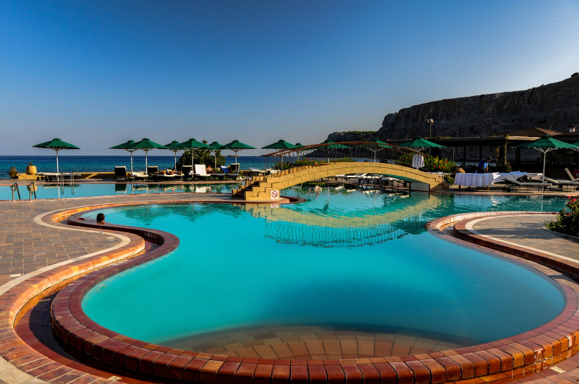 Weitläufige Poollandschaft eines Strandresorts mit Brücke, Liegen und Sonnenschirmen direkt am Meer