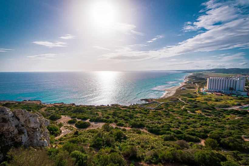 Panoramaaufnahme der Küste von Son Bou auf Menorca mit weiten grünen Dünen, dem langen Sandstrand und einem großen Hotelgebäude am Meer.