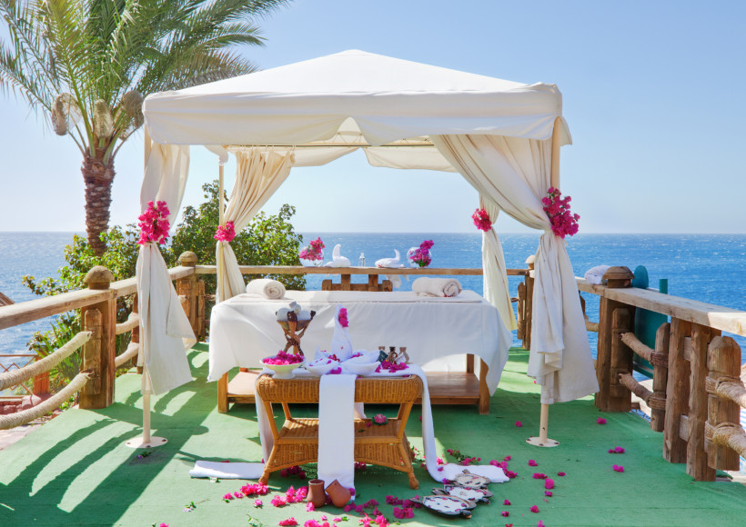 Ägypten Romantisch dekorierte Massageliege unter einem weißen Pavillon mit Vorhängen und pinken Blüten. Der Platz liegt auf einer Holzplattform mit Blick auf das blaue Meer, umgeben von Palmen und Pflanzen.