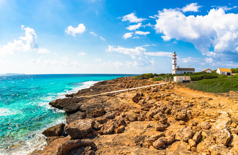 Mallorca - Cap de Ses Salines Eine felsige Küste Mallorcas mit schroffen Klippen und türkisblauem Wasser. Auf einem der Felsen steht ein weißer Leuchtturm mit einem roten Dach, flankiert von kleinen Gebäuden. Im Hintergrund wächst spärliche, typisch mediterrane Vegetation. Die Küstenl