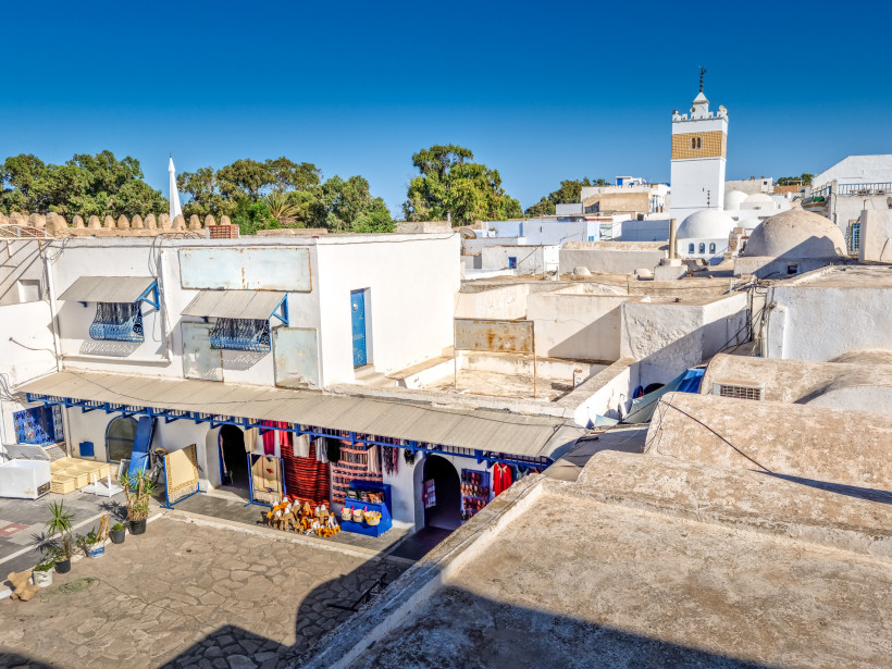 Blick von einer Dachterrasse auf eine traditionelle Medina mit weißen Häusern und flachen Dächern. Im Hintergrund ist ein Minarett zu sehen, das über die Häuser hinausragt. Der blaue Himmel verleiht der Szene eine friedliche und sonnige Atmosphäre.