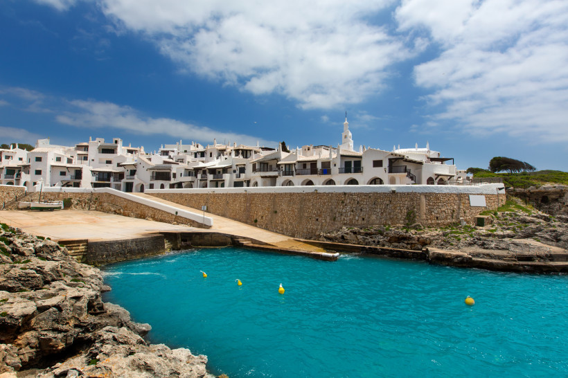 Blick auf Binibeca Vell von der Wasserseite: weiße Häuser mit dunklen Balkonen und Bögen, eine Steinmauer und türkisblaues Meer im Vordergrund.