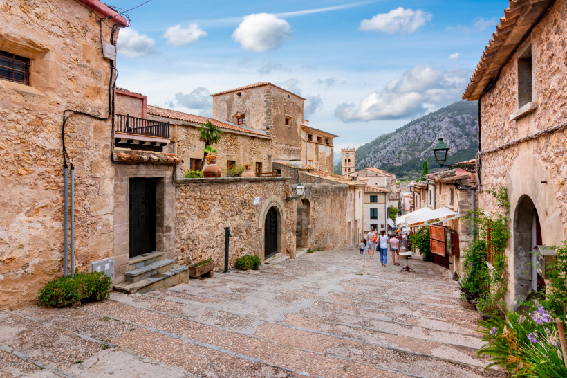 Altstadtgasse in Pollença mit Natursteinhäusern, Treppenweg und Blick auf Kirchturm und Berge
