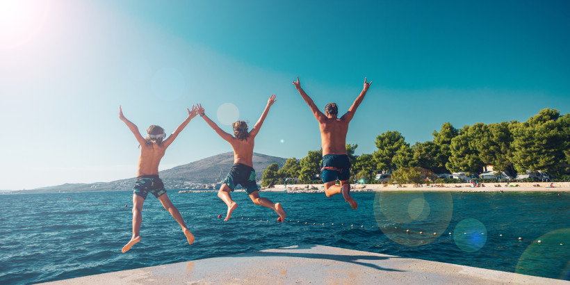 Türkei Drei Jugendliche springen von einem Steg ins Meer, im Hintergrund Strand, Pinien und Wohnmobile