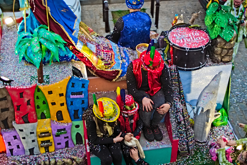 Ein Karnevalswagen mit bunten Dekorationen, Konfetti und fantasievollen Elementen wie einer Palme, einer Stadtfassade und einem Trommelset. Menschen mit glitzernden Hüten und Kostümen sitzen auf dem Wagen. Die gesamte Szene ist von Konfetti bedeckt und st