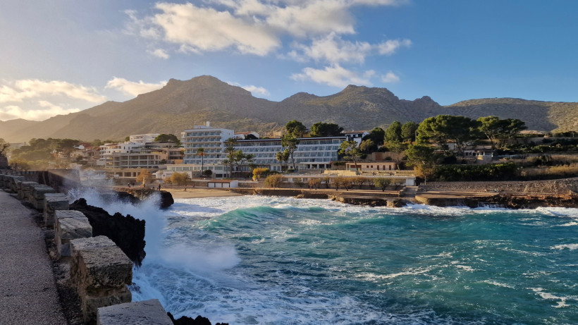 Wellen schlagen an die Uferpromenade von Cala San Vicente, dahinter Hotels und Berglandschaft im Abendlicht.