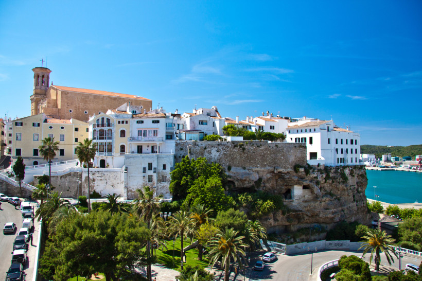 Altstadt von Mahón auf Menorca mit historischen Gebäuden und einer Kirche auf einem Felsplateau. Im Vordergrund Palmen und Blick auf den Naturhafen.
