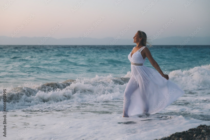 Frau in weißem Kleid steht bei Sonnenuntergang im Meer, sanfte Wellen am Strand und romantische Urlaubsatmosphäre
