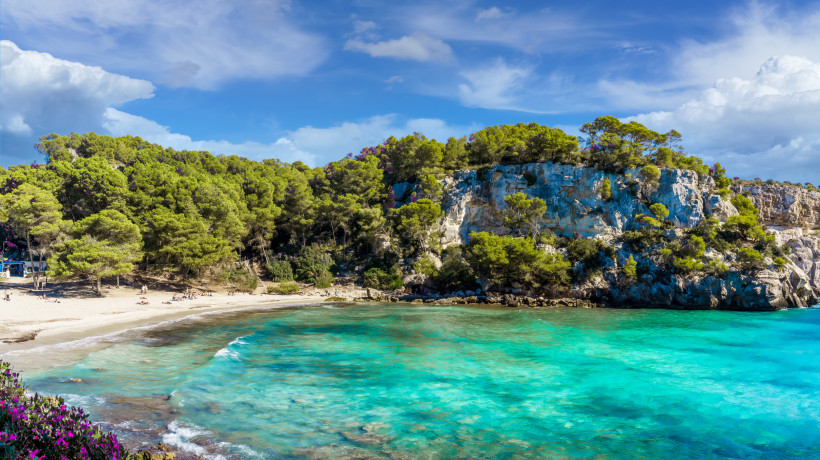 Cala Macarella: Sandstrand, türkisfarbenes Meer und bewaldete Felsküste Bucht Cala Macarella mit Sandstrand, türkisfarbenem Wasser und bewaldeten Felsen unter blauem Himmel