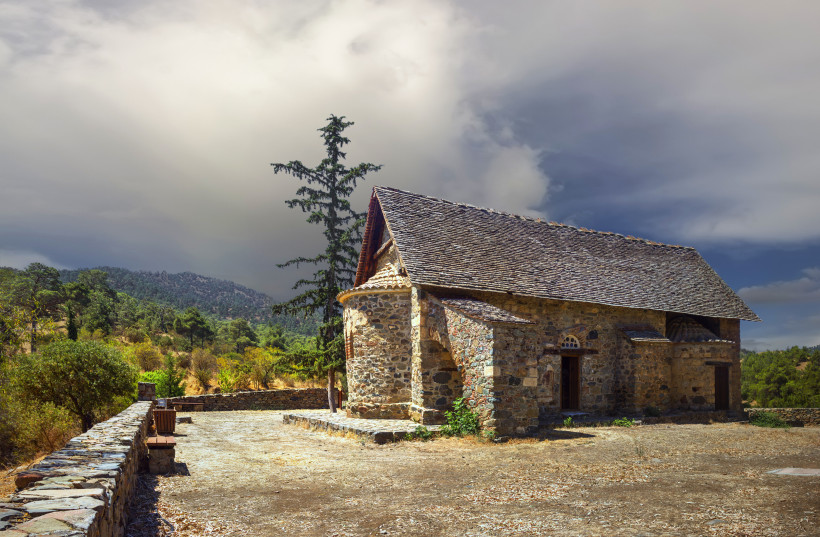 Historische Scheunendachkirche im Troodos-Gebirge auf Zypern: Natursteinbau mit Schieferdach, Zypresse daneben, Berge und dramatischer Himmel.