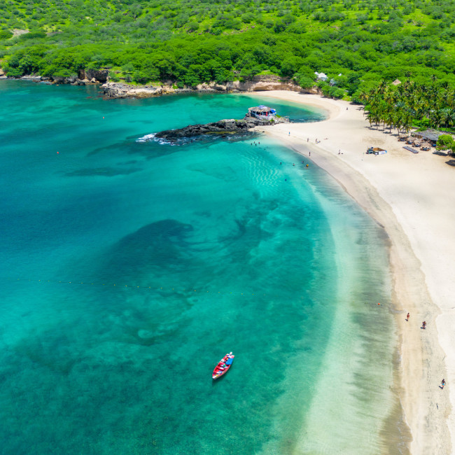 Tropische Badebucht mit türkisblauem Wasser und hellem Sandstrand auf den Kapverden. Am Strand liegen einzelne Badegäste, ein kleines Boot mit Personen treibt im klaren Wasser. Im Hintergrund dichter, grüner Wald und Palmen.