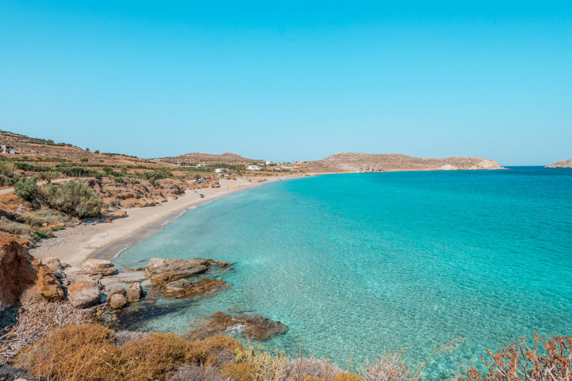 Malerischer Blick auf ein türkisblaues, klares Meer entlang eines Sandstrandes mit Felsen im Vordergrund. Trockenes Hügelland mit spärlicher Vegetation und einige Menschen und Autos in der Ferne. Der Himmel ist strahlend blau und wolkenlos