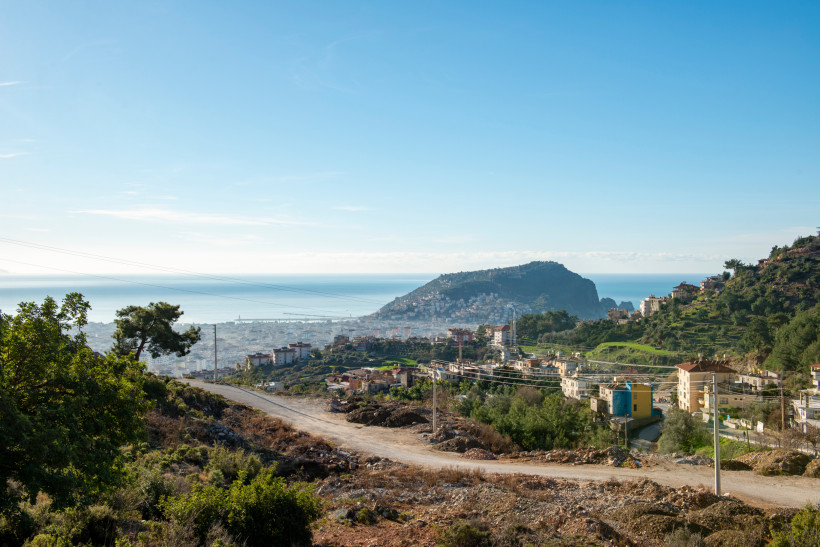 Das Bild zeigt eine Schotterstraße in den Hügeln oberhalb von Alanya. Im Vordergrund liegen verstreute Wohnhäuser und grüne Terrassenhänge. In der Ferne ist die Stadt Alanya mit dem markanten Felsen der Halbinsel und das Mittelmeer zu sehen.