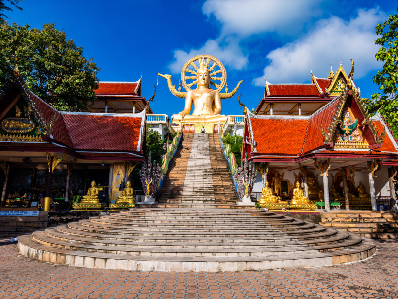Große Buddha-Statue mit Tempelanlage auf Koh Samui – Wat Phra Yai Goldene Buddha-Statue mit rotem Tempeldach auf Koh Samui, Thailand, mit breiter Treppe und blauen Himmel im Hintergrund