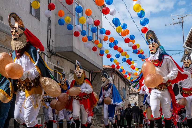 Spanien Straßenszene mit traditionellem Karnevalsumzug. Männer in bunten, folkloristischen Kostümen und kunstvollen Masken mit Hörnern und Bärten marschieren durch eine enge Gasse, über der zahlreiche bunte Luftballons aufgehängt sind. Die Teilnehmer halten große