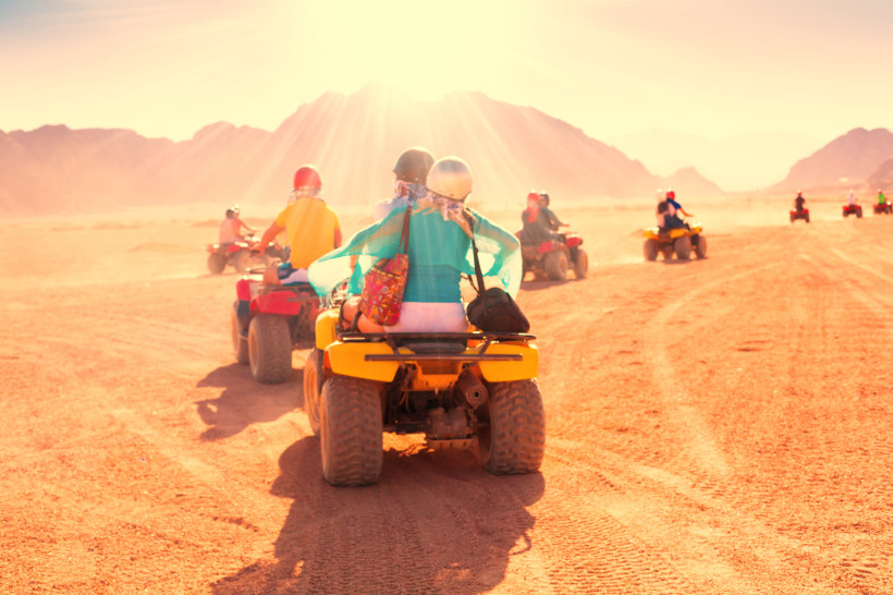 Ägypten Gruppe von Menschen fährt auf Quads durch eine sonnenbeschienene Wüstenlandschaft. Im Vordergrund ein Paar auf einem gelben Quad, das bunte Kleidung und Helme trägt. Im Hintergrund sind weitere Fahrer vor einer Kulisse aus Bergen und staubigem Terrain zu