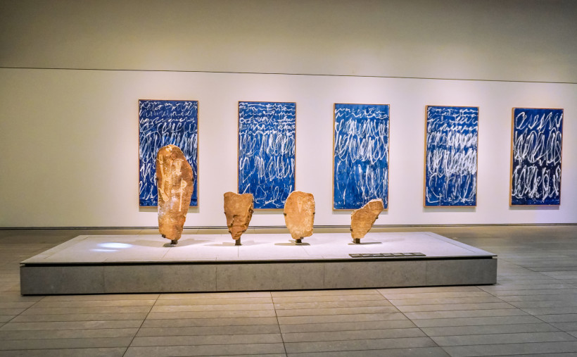 Ausstellungsraum in einem Museum mit alten Steinskulpturen auf einem Podest und fünf blauen Kunstwerken mit weißer Schrift an der Wand