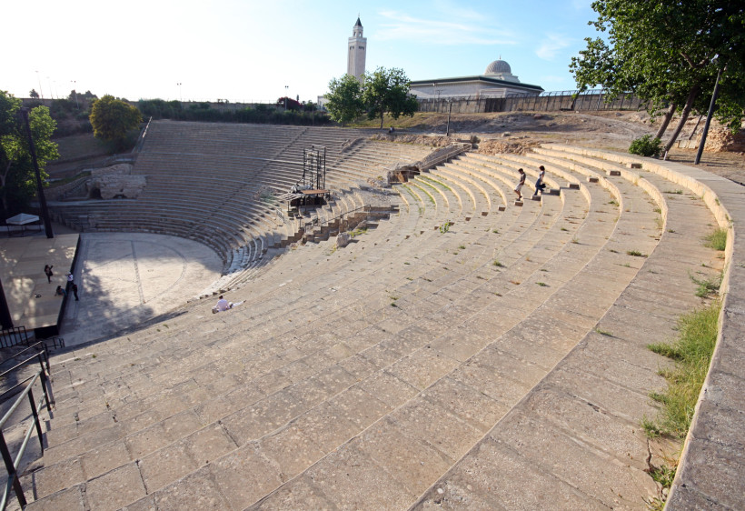 Ein römisches Amphitheater mit steinernen Sitzreihen, die sich halbkreisförmig um eine Arena legen. Im Hintergrund sind Bäume, eine moderne Moschee mit Minarett und Kuppel zu sehen. Einige Menschen sind auf den Stufen unterwegs oder sitzen dort.