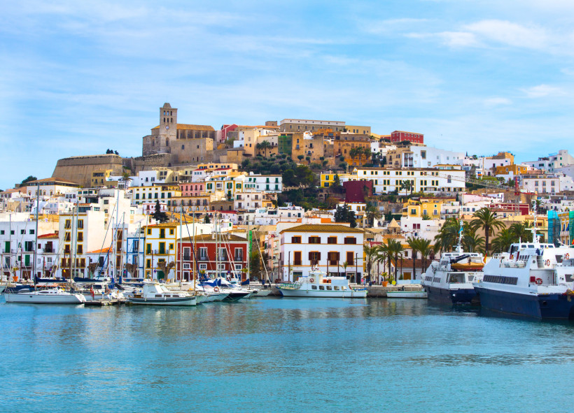 Ibiza Blick auf den Hafen von Ibiza-Stadt mit mehreren weißen Yachten und Segelbooten im Vordergrund. Im Hintergrund erstrecken sich bunte, dicht aneinandergereihte Häuser an einem Hang, überragt von der historischen Altstadt Dalt Vila mit ihrer Festung und der