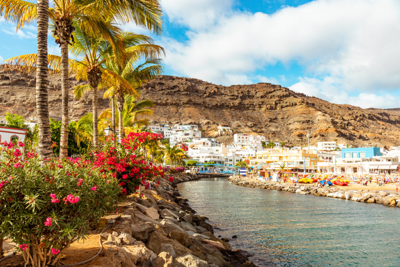 Kanal im Hafen von Puerto de Mogán auf Gran Canaria mit Palmen, blühenden Sträuchern, weißen Häusern und felsigen Bergen im Hintergrund