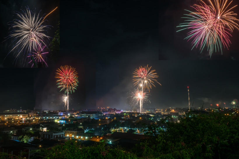 Curaçao Panoramaaufnahme von Willemstad, Curaçao bei Nacht. Mehrere farbenfrohe Feuerwerke explodieren gleichzeitig über der Stadt. Die Häuser sind beleuchtet, Rauch steigt auf, und am Himmel sind rote, grüne und goldene Lichtkronen zu sehen.