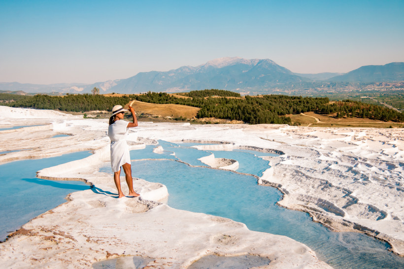 Türkei - Pamukkale Reisende Frau steht auf den weißen Kalkterrassen von Pamukkale und blickt über die türkisfarbenen Thermalbecken mit Bergpanorama im Hintergrund