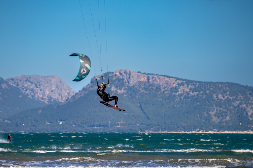 Mallorca Kitesurfer in der Luft über den Wellen, mit Bergen im Hintergrund und windigem Wetter an Mallorcas Küste