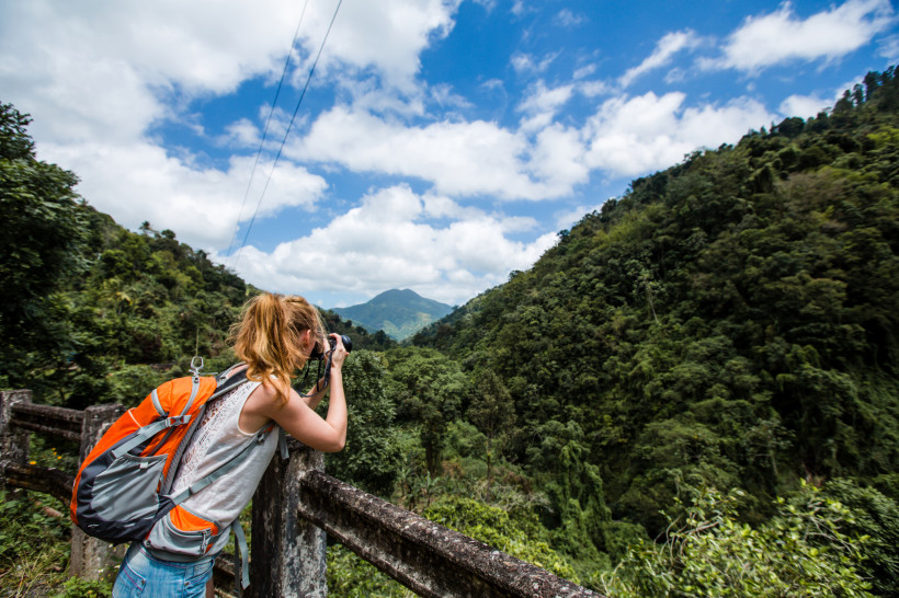 Jamaika Eine junge Frau mit orangem Rucksack steht an einem Geländer und fotografiert mit einer Kamera eine üppig grüne Berglandschaft unter einem blauen Himmel mit weißen Wolken. Die Szene vermittelt ein Gefühl von Abenteuer, Natur und Freiheit.