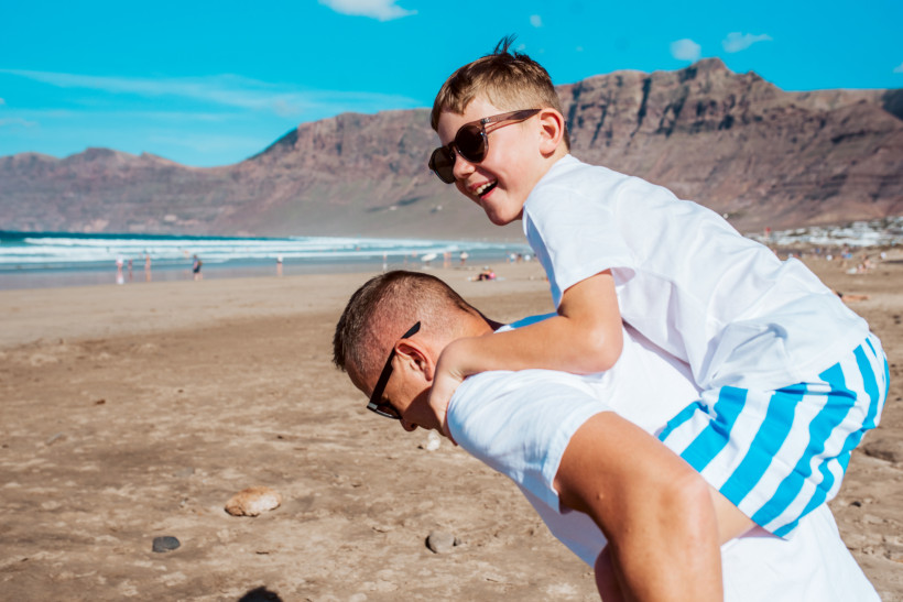Lanzarote Mann trägt lachenden Jungen auf dem Rücken über einen sonnigen Strand. Beide tragen Sonnenbrillen und helle Kleidung, im Hintergrund sind Meer, Berge und andere Strandbesucher zu sehen.