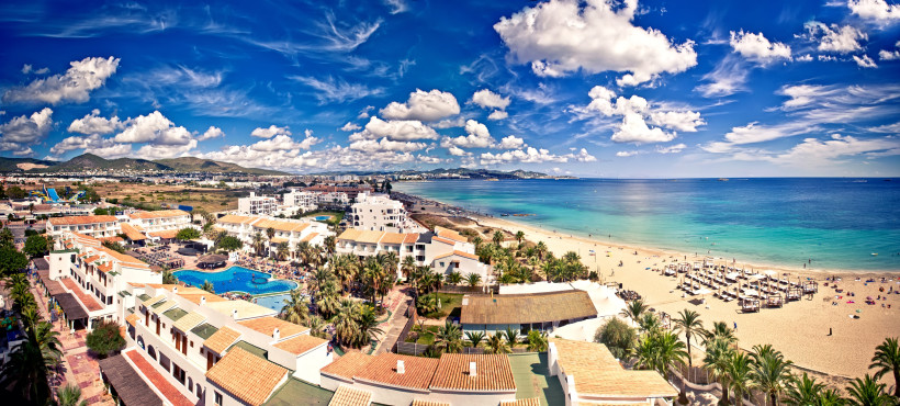 Blick über Playa d'en Bossa auf Sandstrand, türkisfarbenes Meer und Hotelanlagen unter blauem Himmel mit Wolken